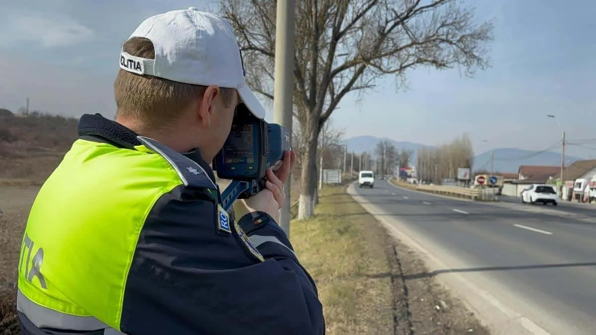 Un bărbat în uniformă albastră, vestă galbenă reflectorizantă și șapcă albă ține un dispozitiv la față, lângă un drum cu vehicule.