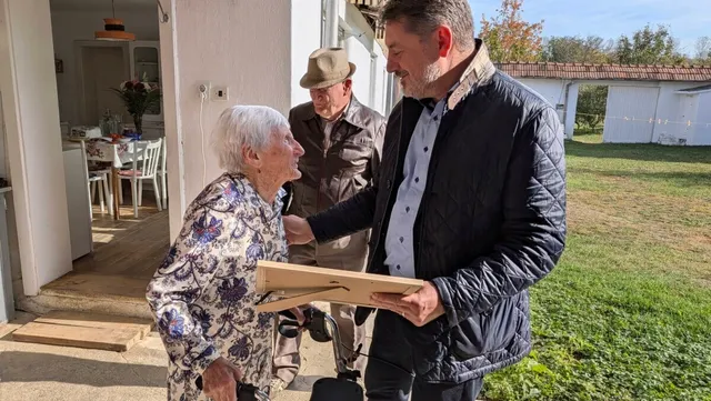 O femeie în vârstă, cu păr alb și bluză cu model floral, stă lângă un bărbat care ține un obiect dreptunghiular; alt bărbat se află în spate.