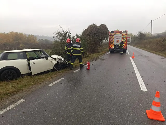 Un autoturism alb, avariat în față, se află lângă șosea; două persoane cu căști stau aproape, iar în spate se vede o autospecială.
