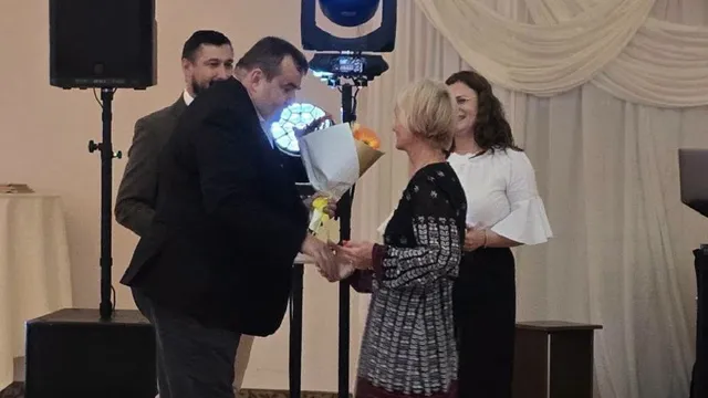 Un bărbat în costum ține un buchet în fața unei femei, iar alte două persoane stau în spatele lor într-o sală decorată.