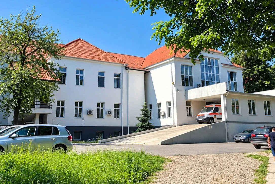 O clădire albă cu acoperiș roșu are o rampă la intrare, o ambulanță și mașini parcate, fiind înconjurată de arbori și iarbă.