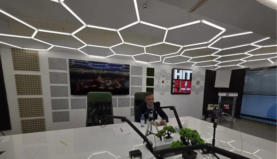 Un bărbat în sacou închis stă la o masă albă cu microfoane și plante verzi, într-o încăpere cu ecrane și lumini geometrice.