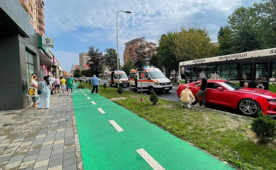 Un autoturism roșu stă pe spațiul verde lângă un autobuz, două ambulanțe, mai mulți pietoni și o pistă verde pentru biciclete.