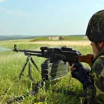 O persoană în haine de camuflaj ține o armă lungă sprijinită pe un suport, lângă o cutie verde, într-un câmp cu iarbă.