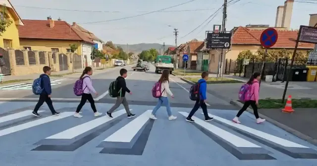 Mai multe persoane traversează șoseaua pe benzi albe și gri în perspectivă, între clădiri, cu vehicule în fundal.