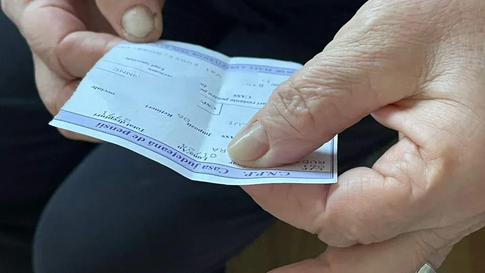 Două mâini țin un carnet mic, deschis, cu pagini albastre și text tipărit, deasupra unei suprafețe întunecate.