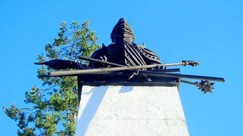 O statuie întunecată, cu o figură în armură și coif, se află deasupra unui piedestal alb, pe fundalul cerului albastru și al unor ramuri.