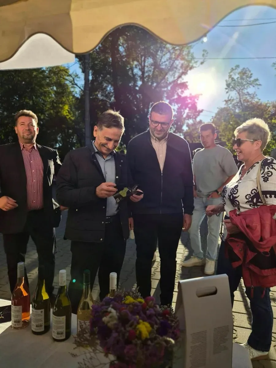 Cinci persoane stau lângă o masă cu sticle de vin și un buchet colorat, sub un cort, în aer liber.