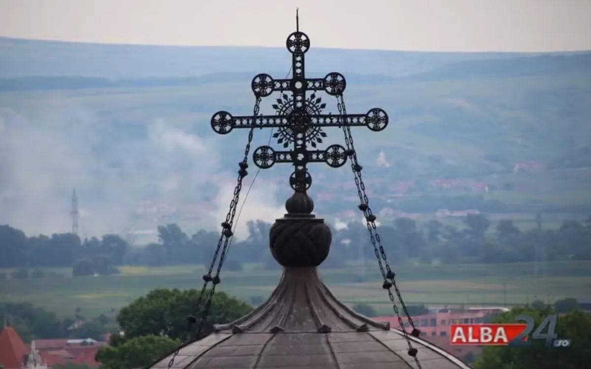 O cruce metalică ornamentată se află deasupra unui acoperiș curbat; în fundal se văd copaci, clădiri și dealuri.