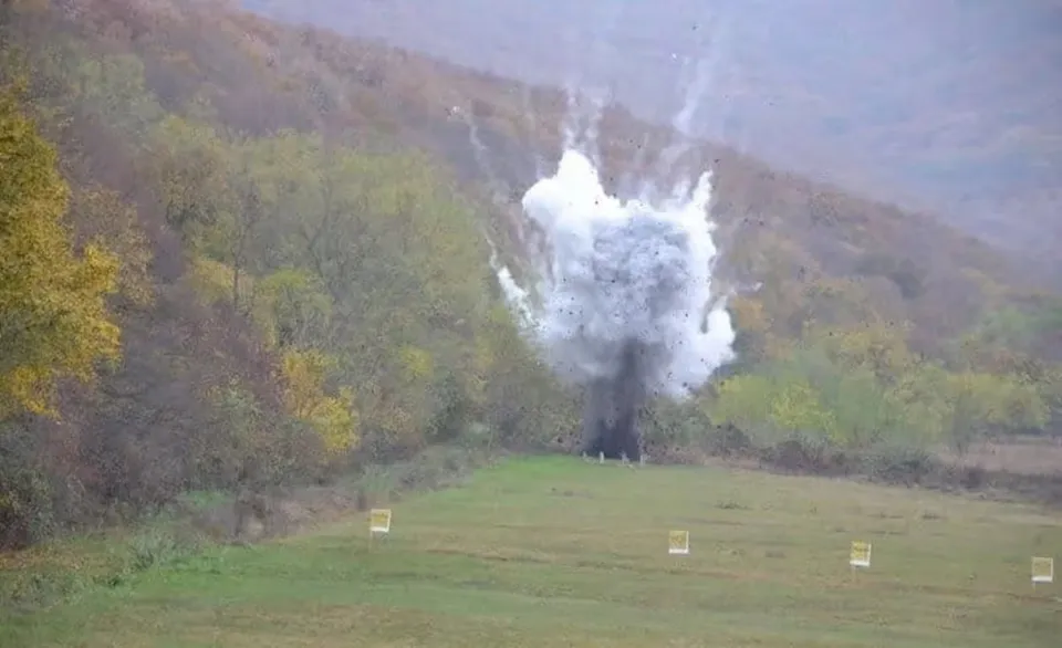 Pe un teren cu iarbă se ridică un nor alb de fum deasupra unei zone întunecate; în față sunt mici panouri dreptunghiulare.