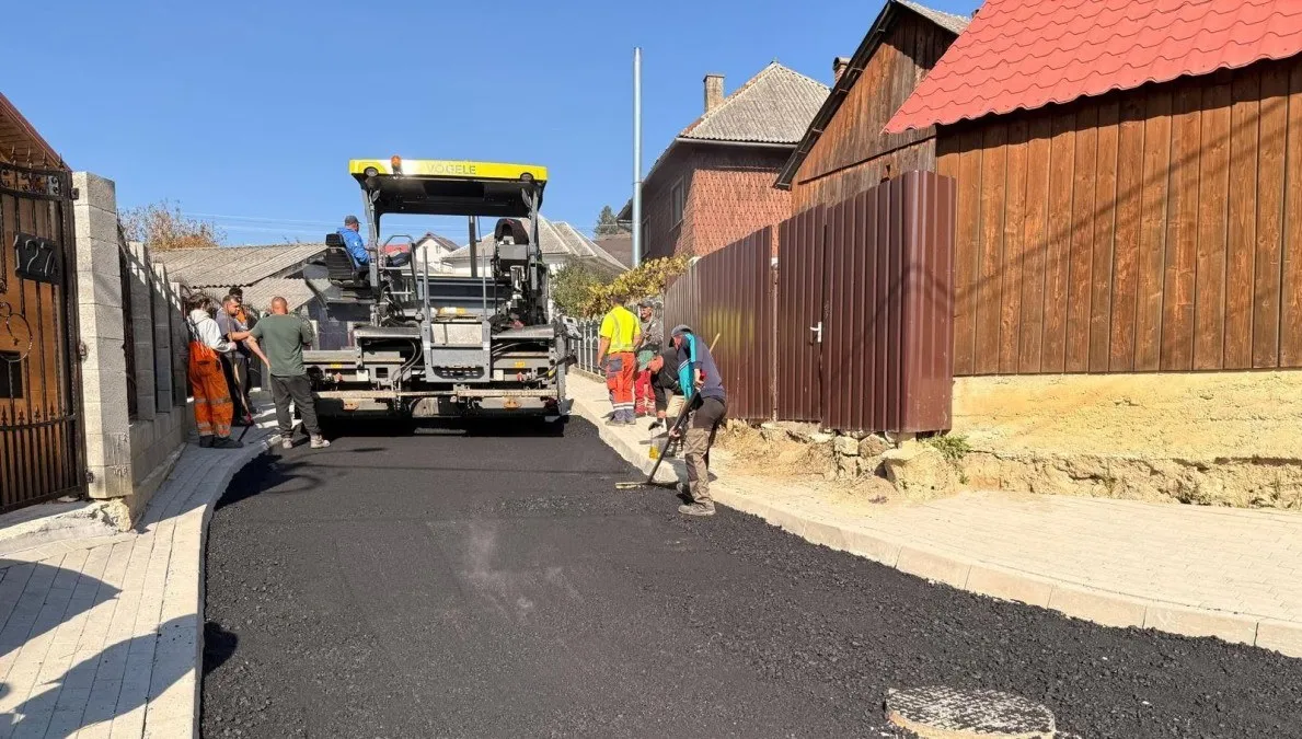 Un utilaj mare se află pe un drum acoperit cu asfalt negru, lângă mai mulți lucrători, garduri, case și trotuare.