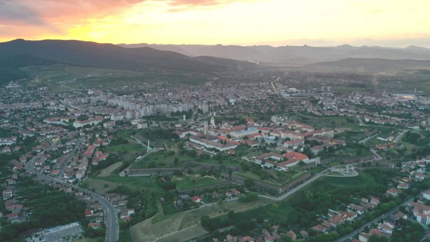 Vedere aeriană asupra unor clădiri grupate în jurul unor terenuri verzi, cu drumuri, cartiere întinse și dealuri la orizont.