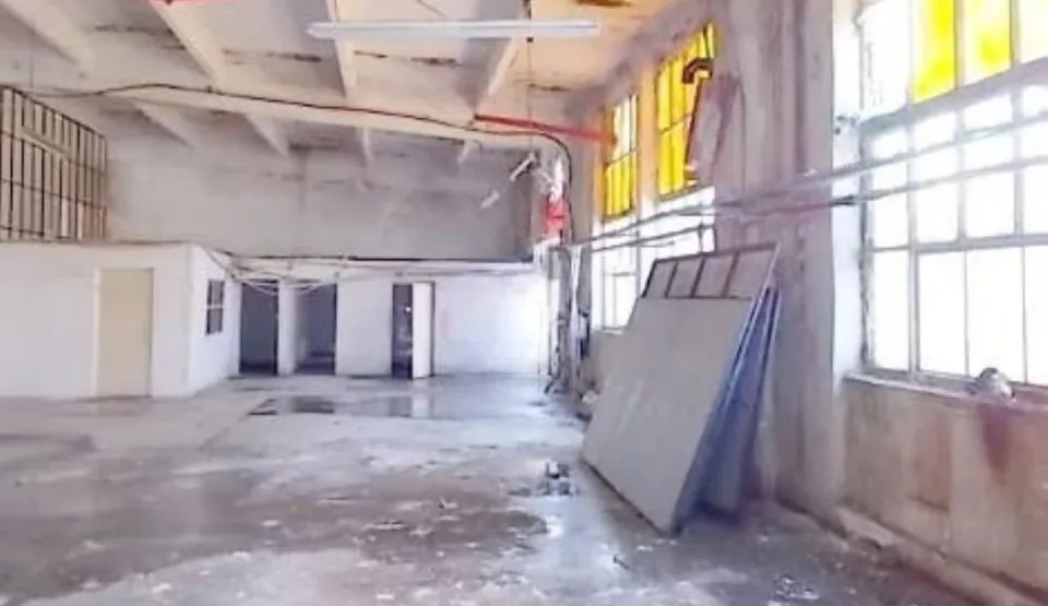 Interiorul unei hale are pereți deteriorați, pardoseală cu resturi, ferestre mari, conducte suspendate și panouri rezemate de un stâlp.