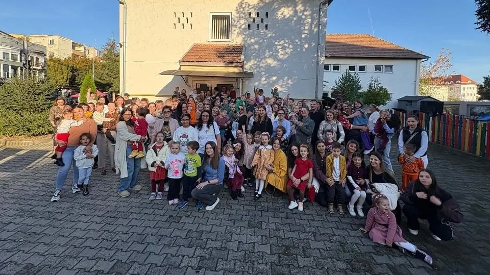 Un grup numeros de adulți și copii stă în picioare și ghemuit în fața unei clădiri, lângă decorațiuni colorate și un gard multicolor.