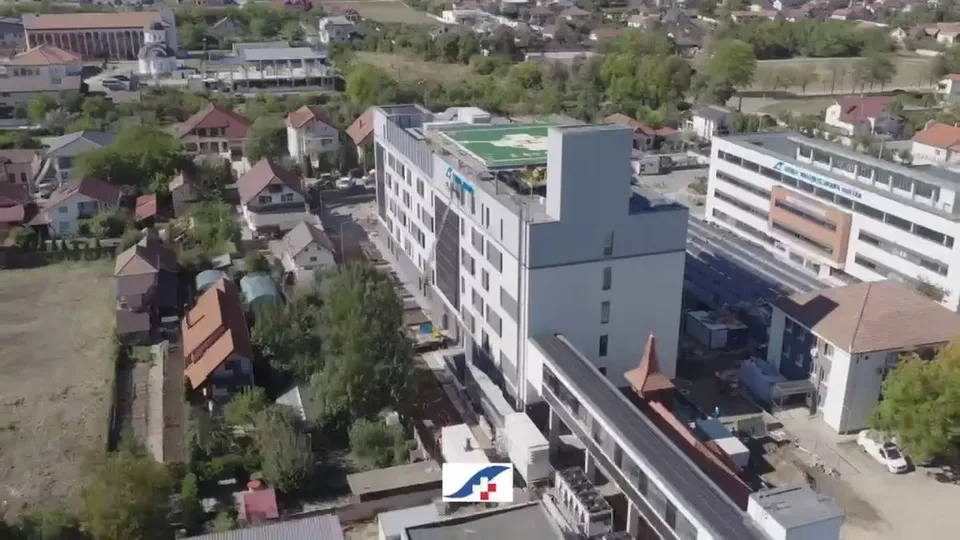 Imagine aeriană cu o clădire modernă cu mai multe etaje, înconjurată de alte clădiri, case, copaci și terenuri.
