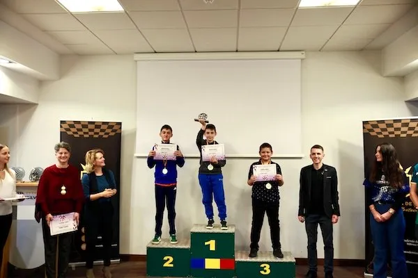 Mai multe persoane stau într-o sală; trei ocupă treptele unui podium numerotat, iar câteva țin diplome sau medalii.