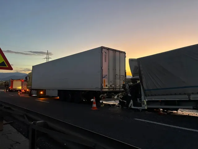 O remorcă albă și un vehicul cu prelată închisă la culoare se află pe șosea, lângă o cabină avariată și un con rutier.