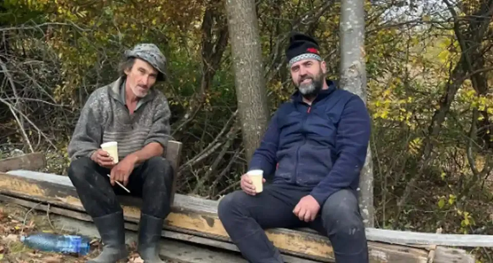 Doi bărbați stau pe scânduri în fața unor copaci; poartă jachete și țin pahare mici în mâini.