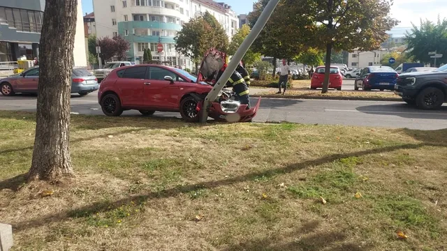 Un autoturism roșu are partea din față avariată, iar un stâlp lung se sprijină oblic peste capotă.