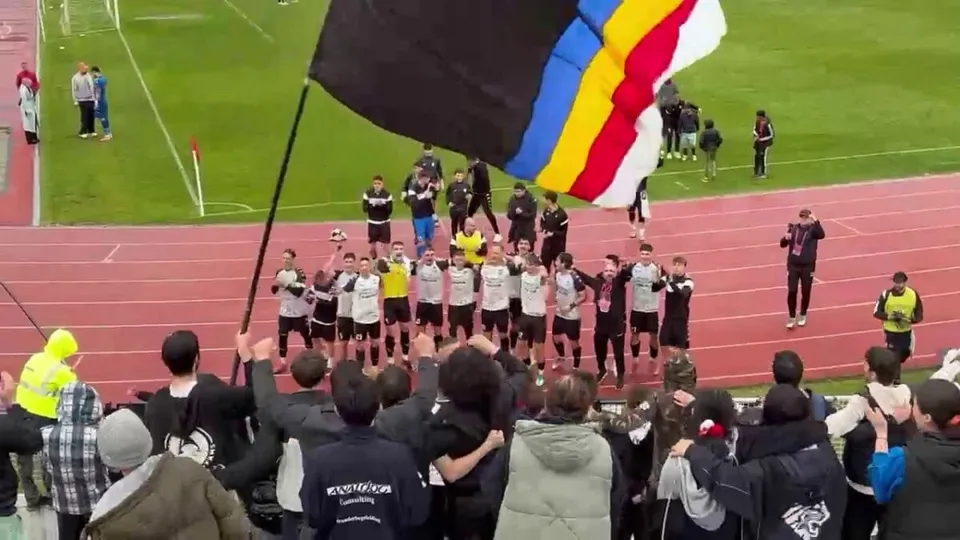 Jucători aliniați pe pista unui stadion au brațele ridicate spre spectatori, sub un steag mare negru, albastru, galben, roșu și alb.