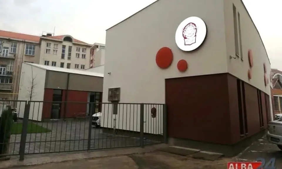 O clădire deschisă la culoare are cercuri roșii și un panou rotund pe fațadă, în spatele unui gard metalic, cu alte clădiri și mașini în jur.