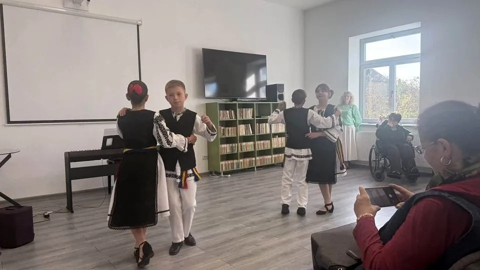 Patru copii în costume populare stau în fața unor persoane așezate, într-o sală cu rafturi de cărți, televizor și fereastră.