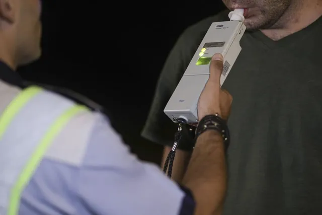 Un bărbat cu vestă reflectorizantă ține un aparat dreptunghiular cu lumini aprinse lângă gura altui bărbat, care suflă într-un muștiuc.