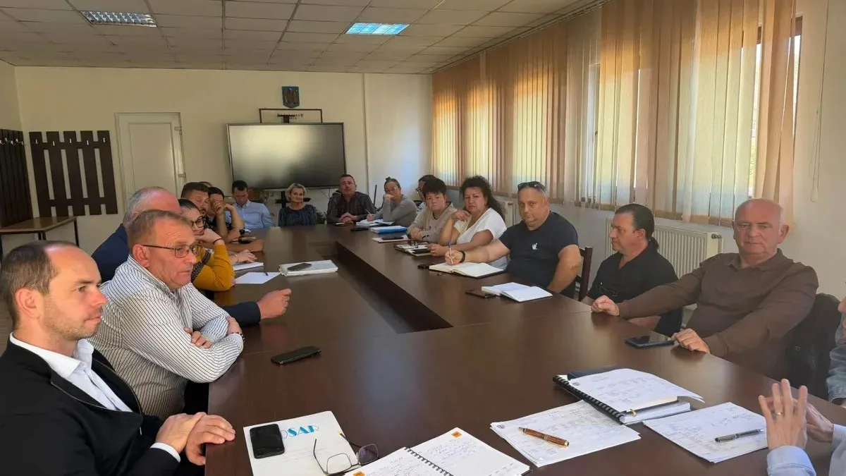 Mai multe persoane stau în jurul unei mese lungi, cu documente și telefoane pe masă; în fundal se văd scaune și jaluzele verticale.