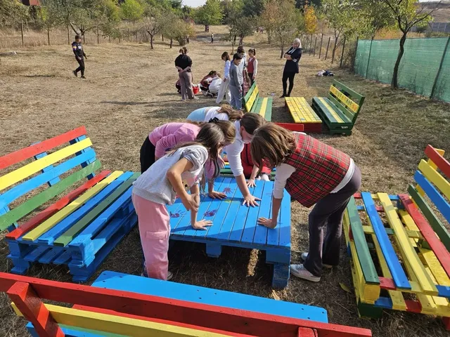 Mai mulți copii se apleacă în jurul unei bănci albastre, printre bănci viu colorate, într-o curte cu iarbă rară și copaci.