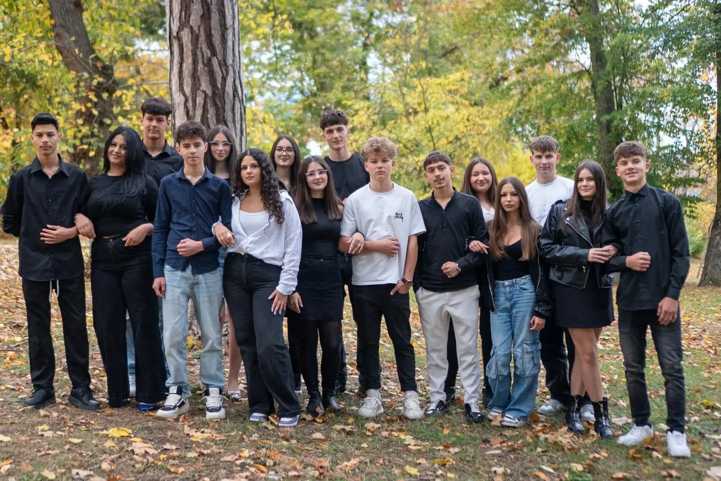 Un grup de tineri în haine casual stă în picioare, aliniat în fața unor copaci, pe un teren acoperit cu frunze.