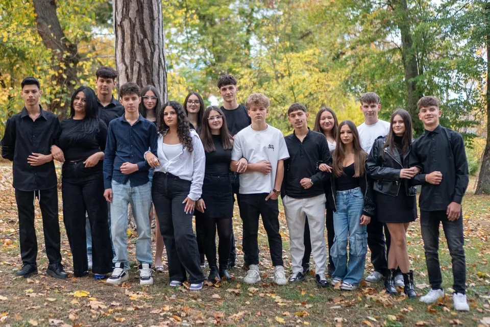 Un grup de tineri în haine casual stă în picioare, aliniat în fața unor copaci, pe un teren acoperit cu frunze.