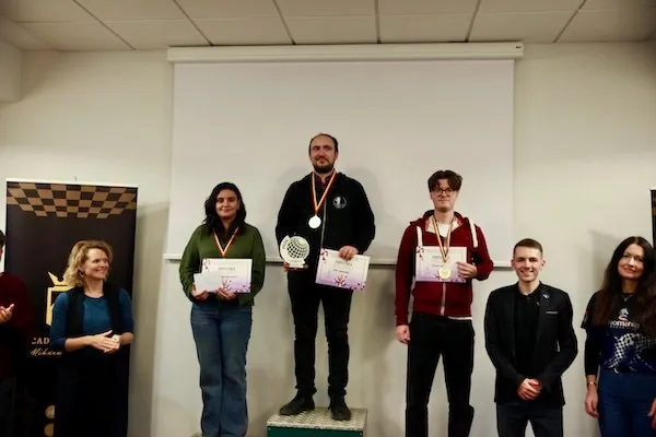 Șase persoane stau în fața unui ecran alb; trei poartă medalii și țin diplome, iar în stânga se vede un panou cu pătrate.