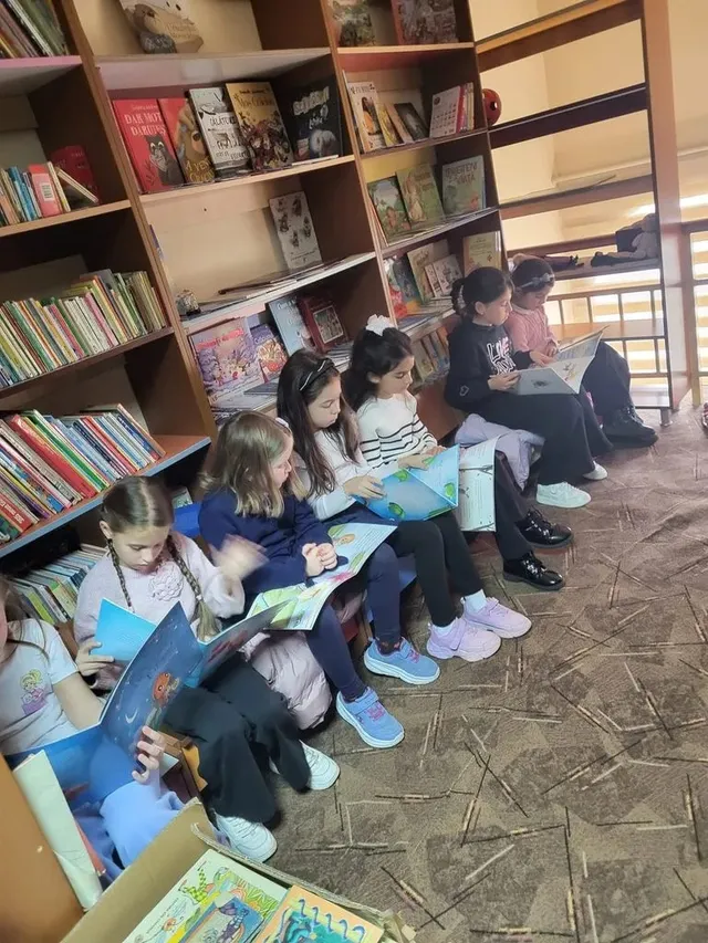 Mai mulți copii stau pe scaune lângă rafturi cu cărți și țin în mâini cărți deschise; pe podea se văd cutii cu cărți.