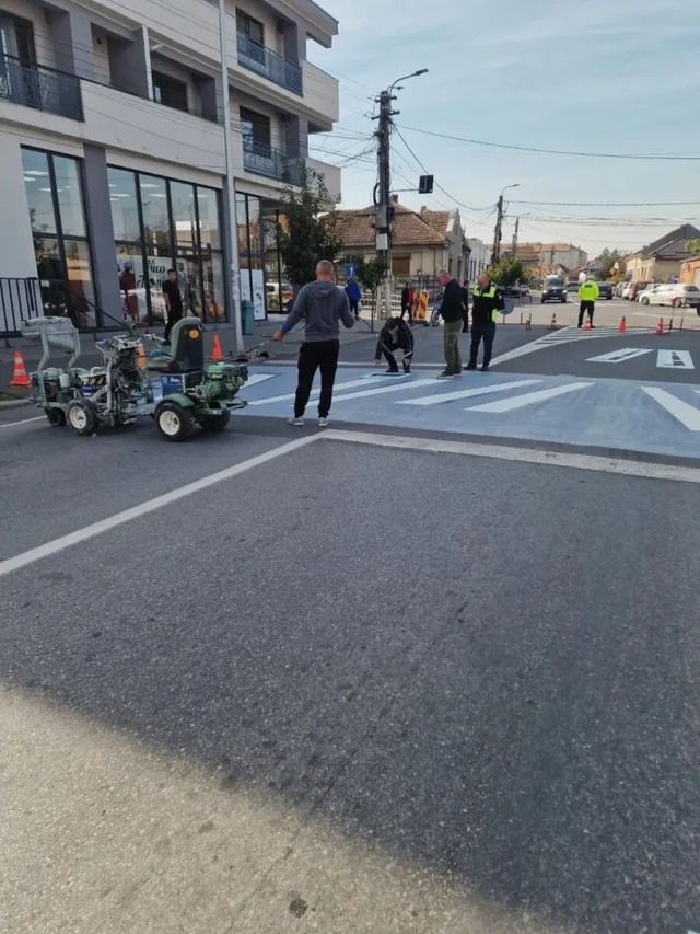 Pe carosabil se vede o trecere cu benzi albe late, mai multe persoane și un utilaj mic lângă conuri portocalii.