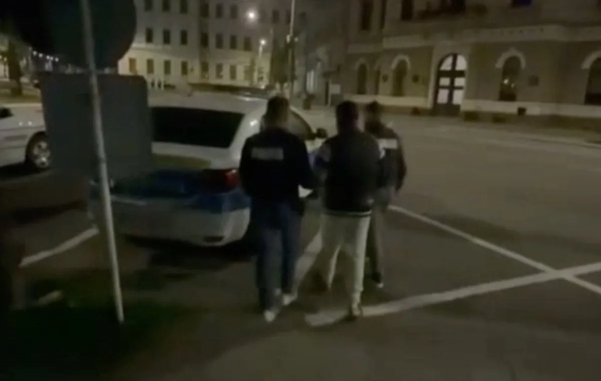 Trei persoane văzute din spate se află lângă un autoturism alb, pe o stradă luminată, între clădiri.