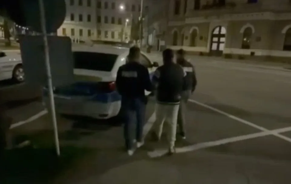 Trei persoane văzute din spate se află lângă un autoturism alb, pe o stradă luminată, între clădiri.