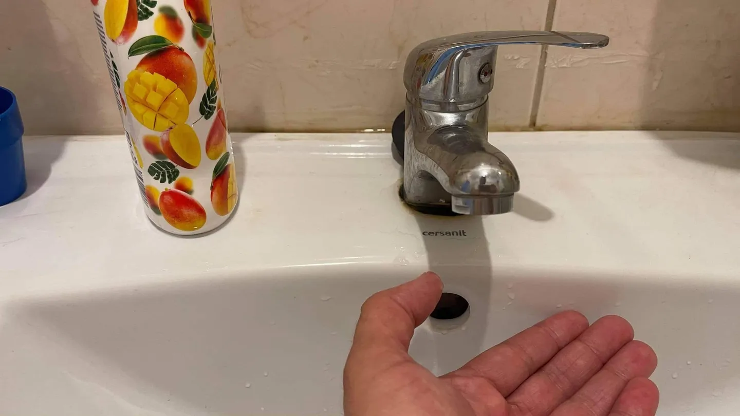 O mână este întinsă sub un robinet, deasupra unei chiuvete albe; lângă robinet se află un recipient cu desene colorate.