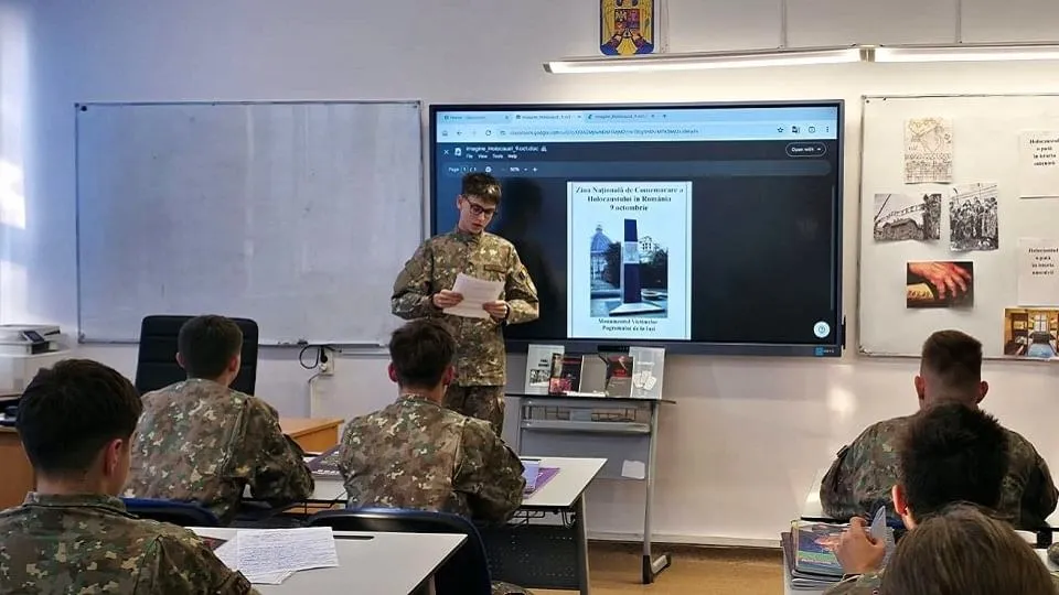 O persoană în uniformă camuflată ține foi în fața unui ecran, iar alte persoane în uniforme camuflate stau la bănci.