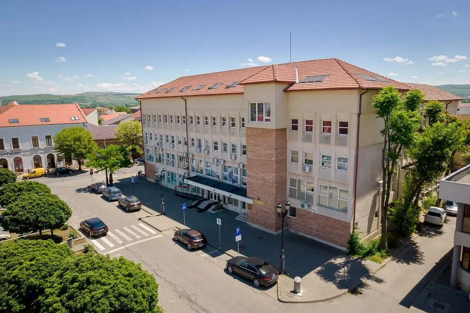 O clădire mare, cu fațadă deschisă și acoperiș roșcat, se află lângă o intersecție cu trecere de pietoni și mai multe mașini.