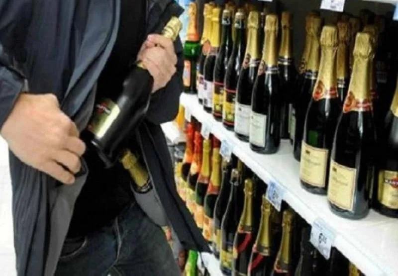O persoană ține o sticlă lângă o haină deschisă, în fața unor rafturi cu sticle.