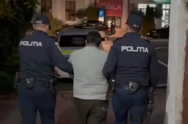 Doi oameni în uniforme închise la culoare merg lângă o persoană cu jachetă deschisă la culoare, văzuți din spate, lângă o mașină.
