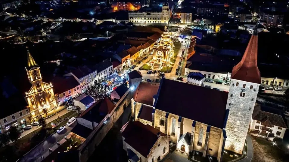 O vedere aeriană nocturnă arată o biserică, clădiri, străzi și șiruri de standuri luminate într-o piață.
