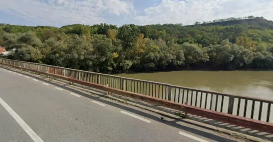 Un pod rutier cu balustradă metalică traversează un râu, iar pe malul opus se văd copaci și un deal împădurit.