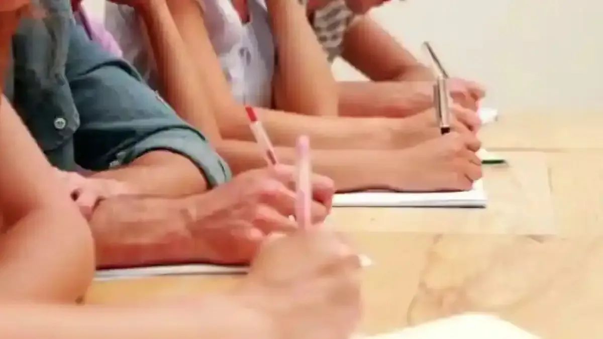 Mai multe persoane așezate la o masă țin pixuri deasupra unor foi albe; în prim-plan se văd mâini și o foaie.