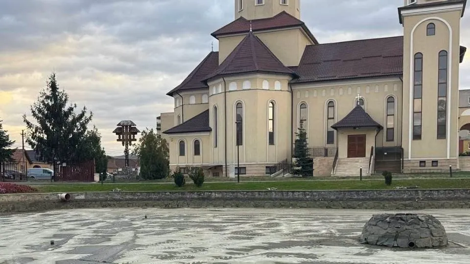 În prim-plan se vede o suprafață de apă cu o piatră mare, iar în spate se află o clădire cu turnuri, copaci și un cer înnorat.