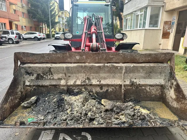În fața unui utilaj roșu, o cupă metalică conține grămezi de material cenușiu și pietre; în fundal se văd clădiri și mașini parcate.