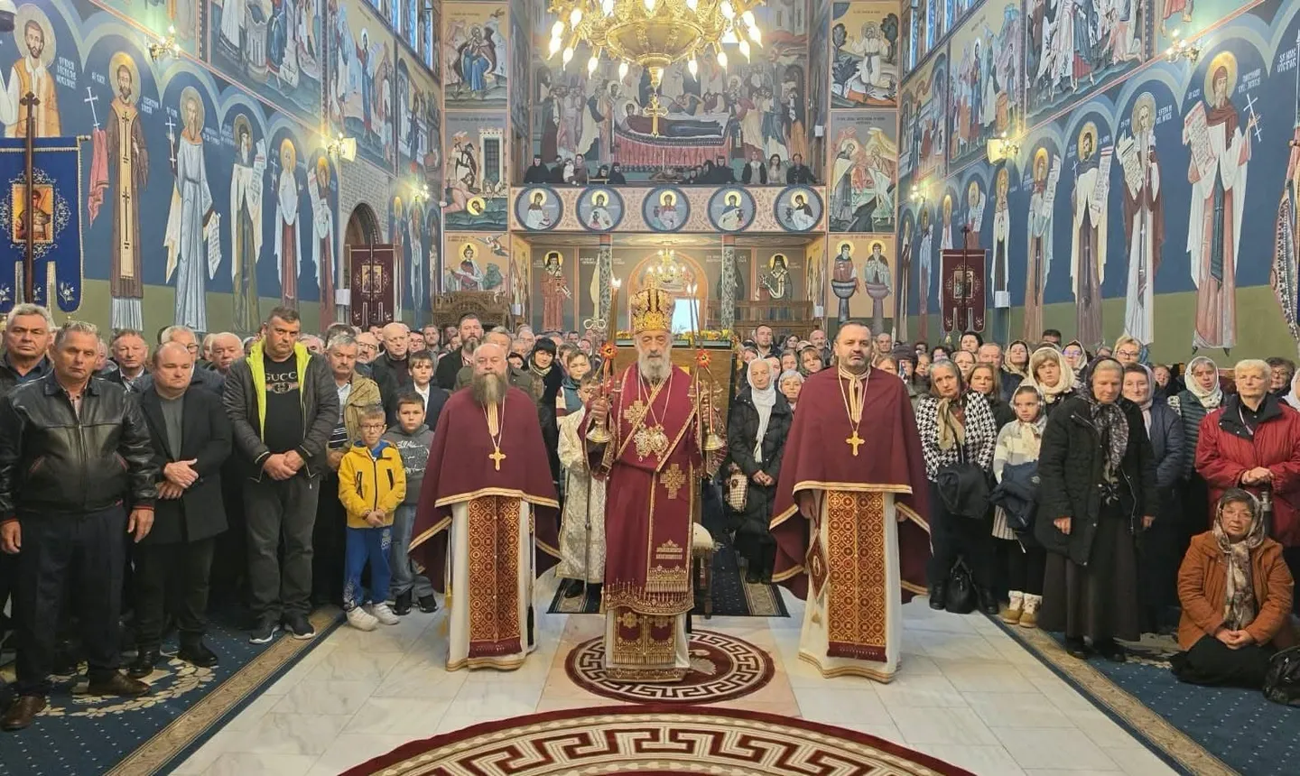Trei clerici în veșminte roșii stau în fața unui grup de oameni, într-un interior cu pereți pictați și candelabre.