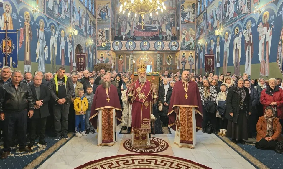 Trei clerici în veșminte roșii stau în fața unui grup de oameni, într-un interior cu pereți pictați și candelabre.