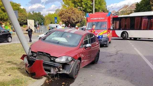 Un autoturism roșu cu partea frontală avariată se află pe carosabil, lângă o autospecială roșie și un autobuz alb.