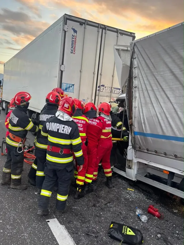 Mai mulți salvatori în uniforme roșii și negre stau între două vehicule mari, lângă fragmente împrăștiate pe carosabil.